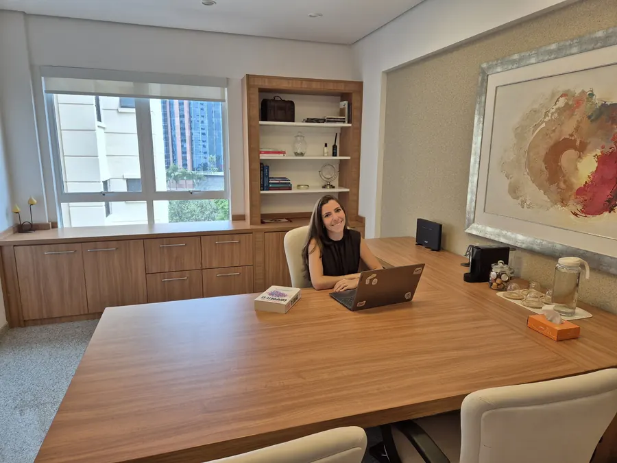 Carolina Cervone na sala de atendimento do consultório, com mesa de madeira, estante e quadro